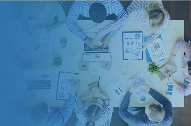 Overhead view of a group of professionals seated around a table reviewing charts and data on laptops and printed reports, representing collaborative planning and analysis.
